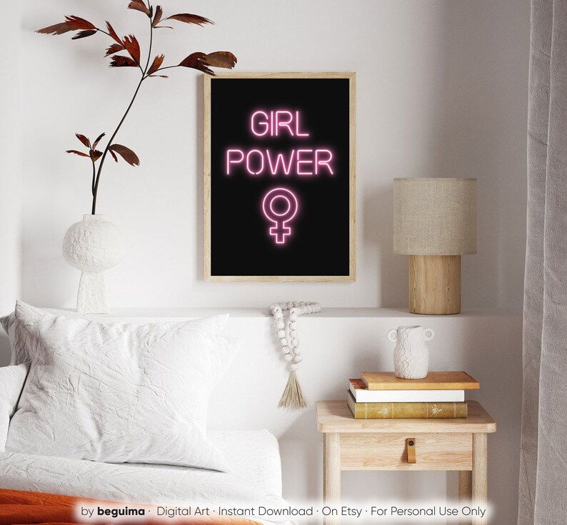 May include: A framed art print featuring the words "GIRL POWER" in vibrant pink neon, and the female symbol, set against a black backdrop. The print is in a light wood frame. The artwork is displayed on a white shelf with decorative items.