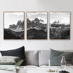 May include: Set of three framed black and white mountain landscape prints. The images depict rugged peaks with clouds and forests. The frames are a warm brown color, and the overall aesthetic is minimalist and natural.