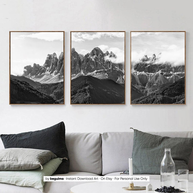 May include: Set of three framed black and white mountain landscape prints. The images depict rugged peaks with clouds and forests. The frames are a warm brown color, and the overall aesthetic is minimalist and natural.