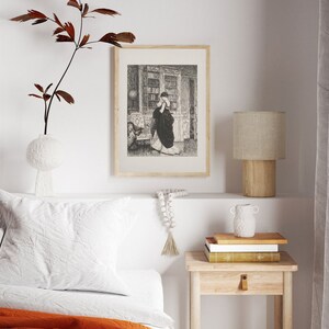 Peut inclure: Une impression encadr&eacute;e en noir et blanc d'une femme dans une biblioth&egrave;que. L'impression est affich&eacute;e au-dessus d'une petite table de chevet en bois avec une lampe, des livres et un vase d&eacute;coratif. Un oreiller blanc et une couverture orange sont visibles.