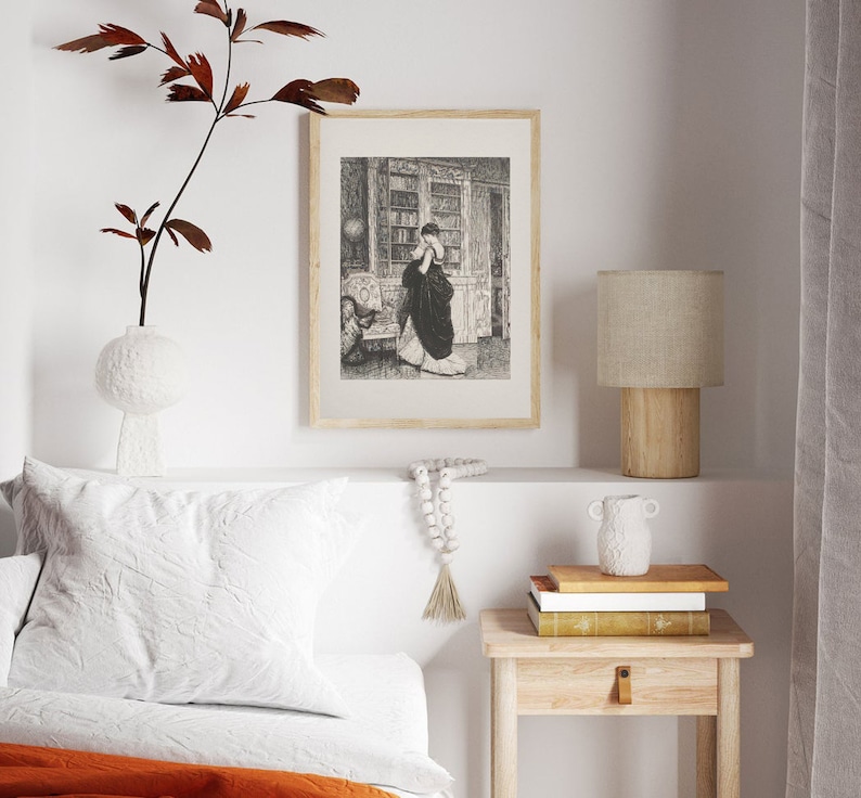 Peut inclure: Une impression encadr&eacute;e en noir et blanc d'une femme dans une biblioth&egrave;que. L'impression est affich&eacute;e au-dessus d'une petite table de chevet en bois avec une lampe, des livres et un vase d&eacute;coratif. Un oreiller blanc et une couverture orange sont visibles.