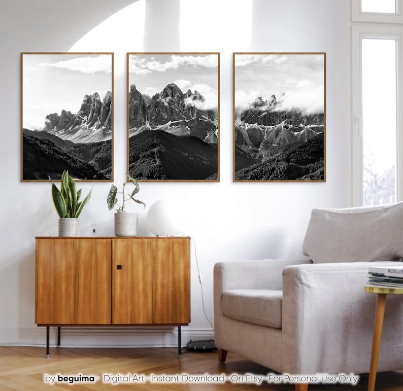 May include: Three framed black and white mountain landscape prints. The artwork features dramatic peaks, clouds, and valleys. The frames are a warm brown color, and the prints are displayed above a wooden cabinet.