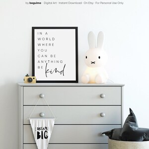 May include: A black and white printable wall art print with the text "In a world where you can be anything be kind". The print is displayed on a gray dresser with a white bunny lamp, a black and white pennant banner that says "Dream Big", and a black pillow in a woven basket.