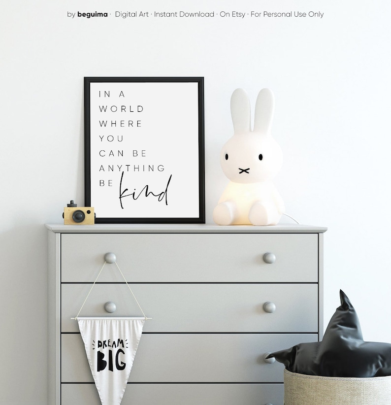 May include: A black and white printable wall art print with the text "In a world where you can be anything be kind". The print is displayed on a gray dresser with a white bunny lamp, a black and white pennant banner that says "Dream Big", and a black pillow in a woven basket.