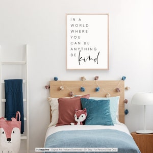 May include: A framed print with a white background and black text that reads "In a world where you can be anything be kind". The print is hanging on a white wall above a wooden bed with blue and red pillows.