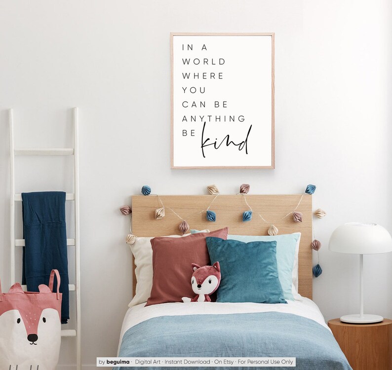 May include: A framed print with a white background and black text that reads "In a world where you can be anything be kind". The print is hanging on a white wall above a wooden bed with blue and red pillows.