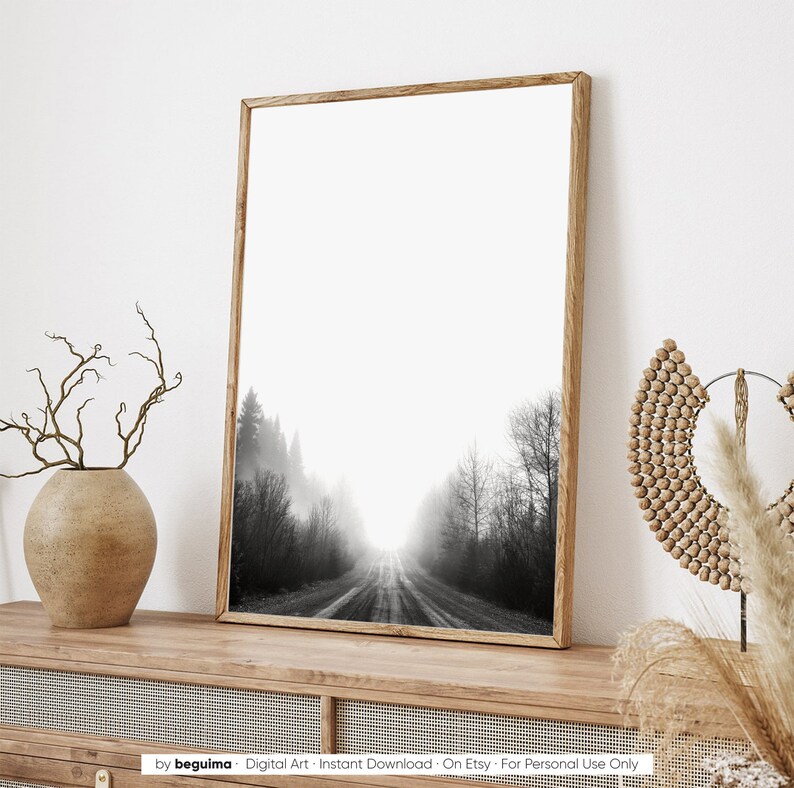 May include: Black and white framed art print featuring a misty forest road. The artwork is in a wooden frame and is displayed on a wooden cabinet. The print depicts a road leading into a bright, foggy area, with trees on either side.