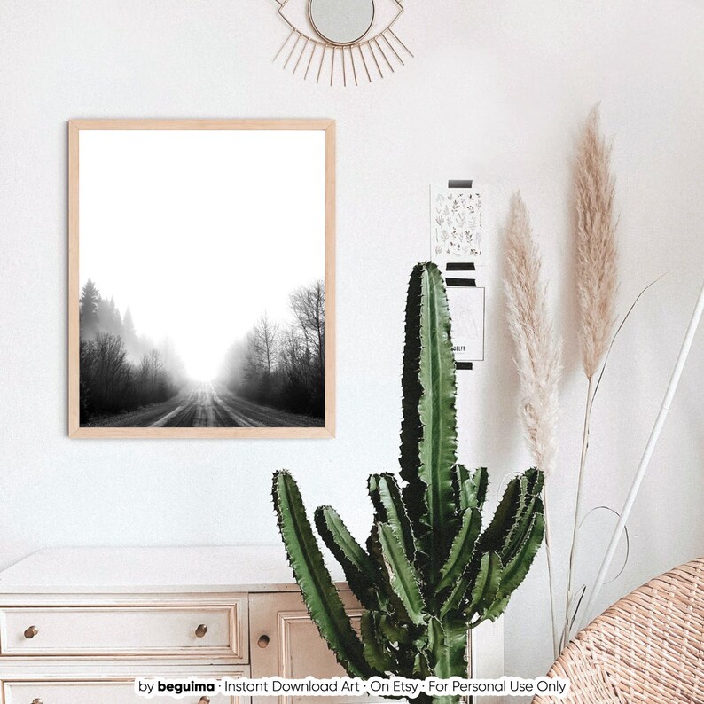 May include: A framed black and white landscape print depicts a road disappearing into a misty forest. The artwork is in a light wood frame, displayed on a white wall. A tall cactus and decorative elements are in the foreground.