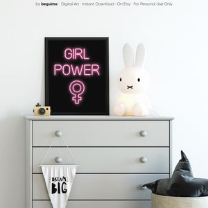 May include: A black framed print with pink neon text reading "GIRL POWER" and a female symbol. A white bunny lamp and a wooden camera are on a grey chest of drawers. A banner below says "DREAM BIG".