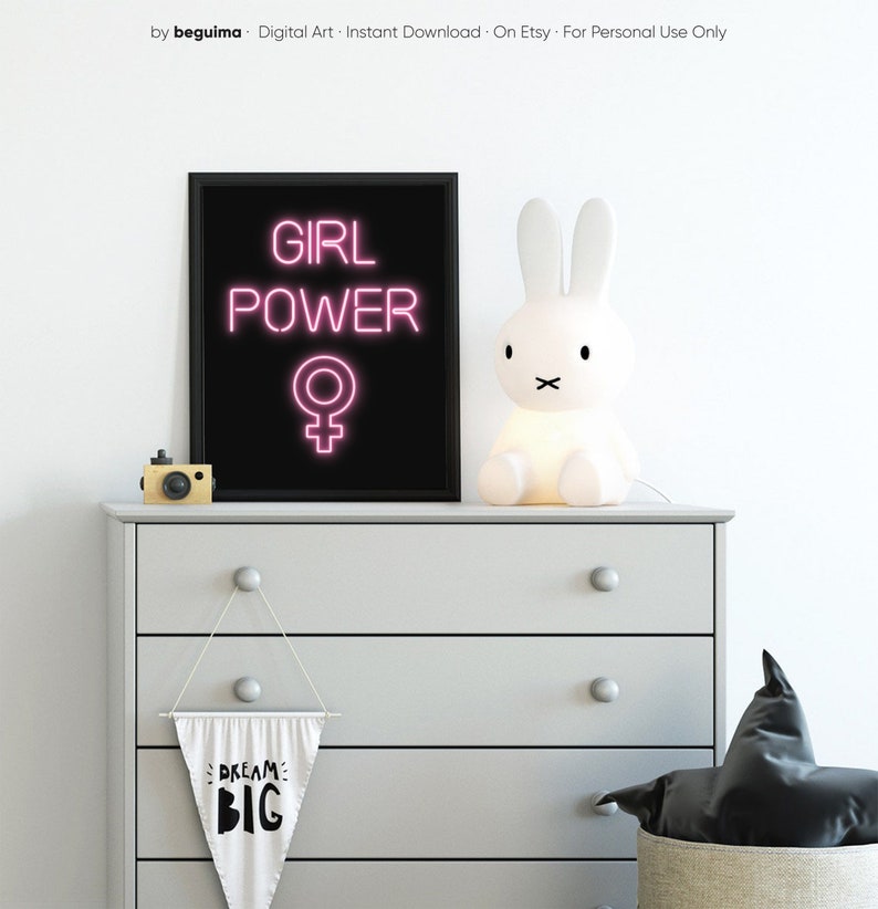 May include: A black framed print with pink neon text reading "GIRL POWER" and a female symbol. A white bunny lamp and a wooden camera are on a grey chest of drawers. A banner below says "DREAM BIG".