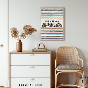 May include: A framed art print with the text "WE ARE ALL DIFFERENT AND THAT'S BEAUTIFUL" over a background of colourful horizontal stripes. The print hangs above a white chest of drawers, a vase with dried plants, and a woven chair.