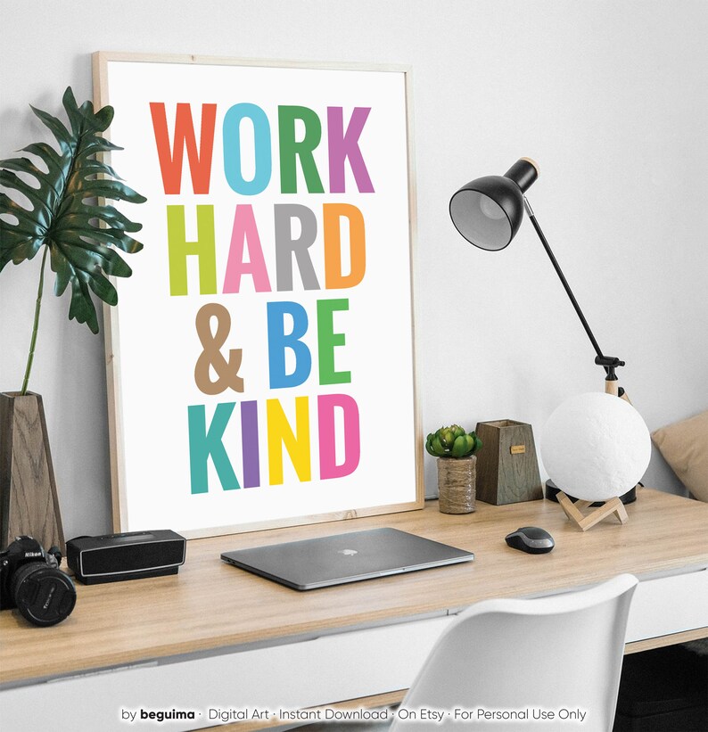 May include: A framed motivational poster with the text "WORK HARD & BE KIND" in colorful letters. The poster is hanging on a white wall above a desk with a laptop, a lamp, and a moon lamp.