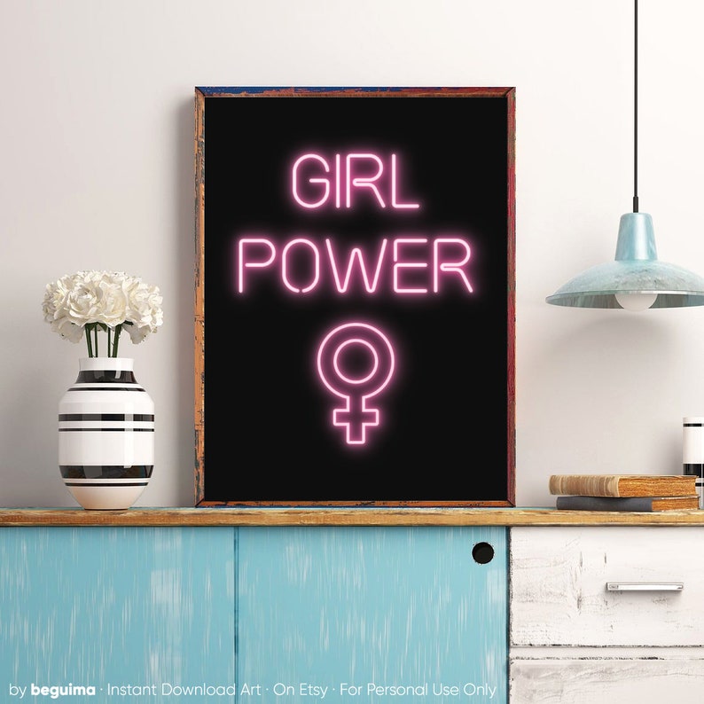 May include: A framed black print featuring the words "GIRL POWER" in pink neon lettering and the female symbol. The artwork is displayed on a blue cabinet. A vase of white flowers is also visible.