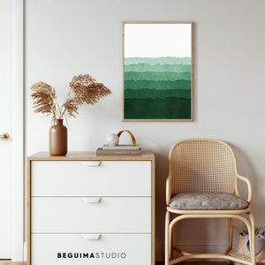 May include: A framed watercolor print with a gradient of green hues, from light to dark, hangs on a white wall. A wooden dresser with three white drawers and the text "BEGUIMASTUDIO" is in front of the wall. A woven chair and decorative items complete the scene.