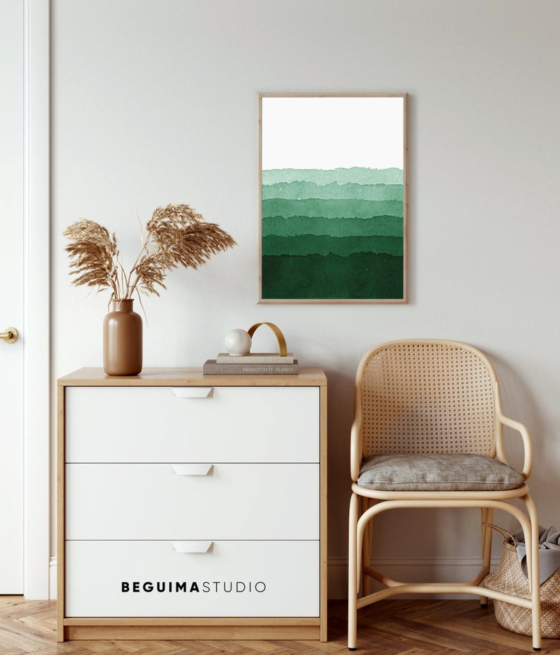 May include: A framed watercolor print with a gradient of green hues, from light to dark, hangs on a white wall. A wooden dresser with three white drawers and the text "BEGUIMASTUDIO" is in front of the wall. A woven chair and decorative items complete the scene.