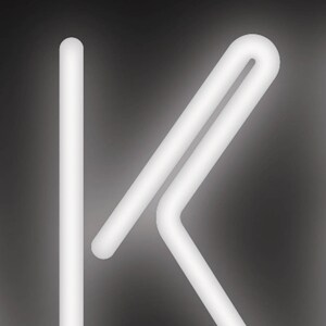 May include: A white, glowing neon light in the shape of the letter "K" against a dark gray background. The "K" is formed by a straight vertical line and a connected, angled design.