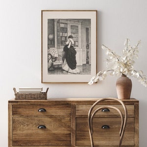 Peut inclure: Une impression encadr&eacute;e en noir et blanc d'une femme en robe longue dans une biblioth&egrave;que. L'impression est affich&eacute;e au-dessus d'une commode en bois avec un panier en osier et un vase avec des fleurs blanches. Une chaise en bois est devant.