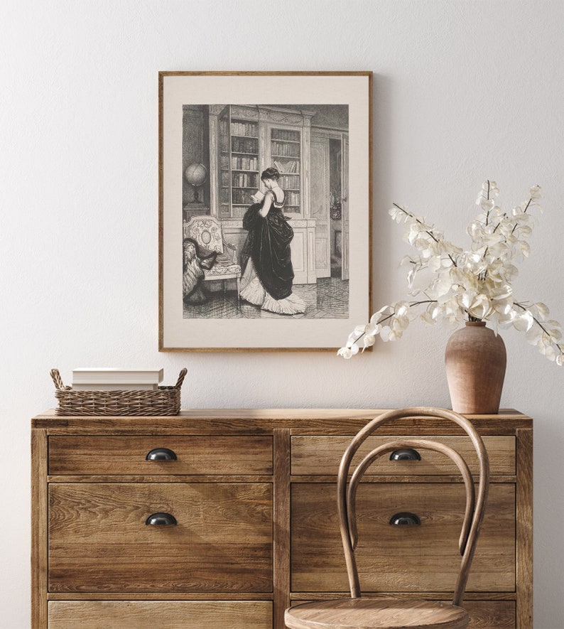 Peut inclure: Une impression encadr&eacute;e en noir et blanc d'une femme en robe longue dans une biblioth&egrave;que. L'impression est affich&eacute;e au-dessus d'une commode en bois avec un panier en osier et un vase avec des fleurs blanches. Une chaise en bois est devant.