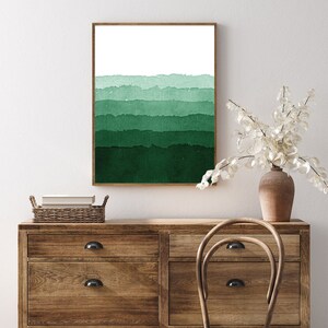 May include: A framed watercolor print featuring horizontal bands of varying shades of green, transitioning from light to dark. The artwork is displayed on a white wall above a wooden dresser. The print has a brown frame.