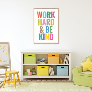 May include: A framed print with the text "WORK HARD & BE KIND" in colorful letters on a white background. The print is hanging on a white wall in a room with a bookshelf and a green armchair.