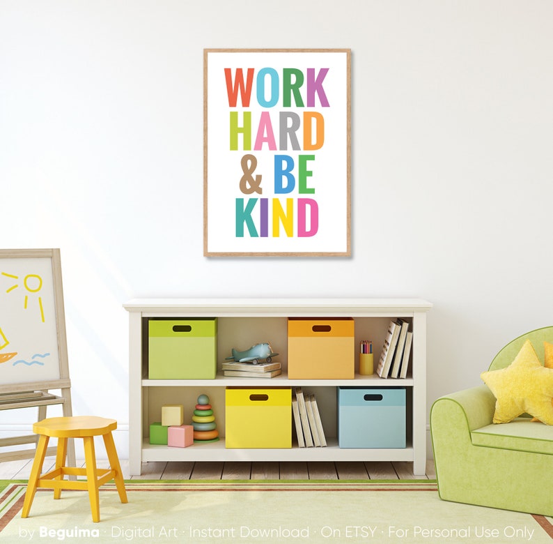 May include: A framed print with the text "WORK HARD & BE KIND" in colorful letters on a white background. The print is hanging on a white wall in a room with a bookshelf and a green armchair.