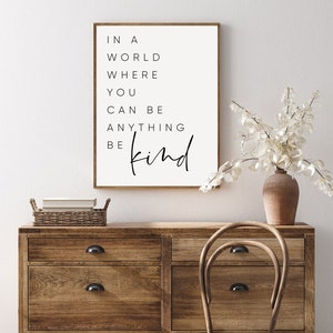 May include: A white framed print with black text that reads "In a world where you can be anything be kind". The print is hanging on a white wall above a wooden dresser with a wicker basket and a wooden chair.