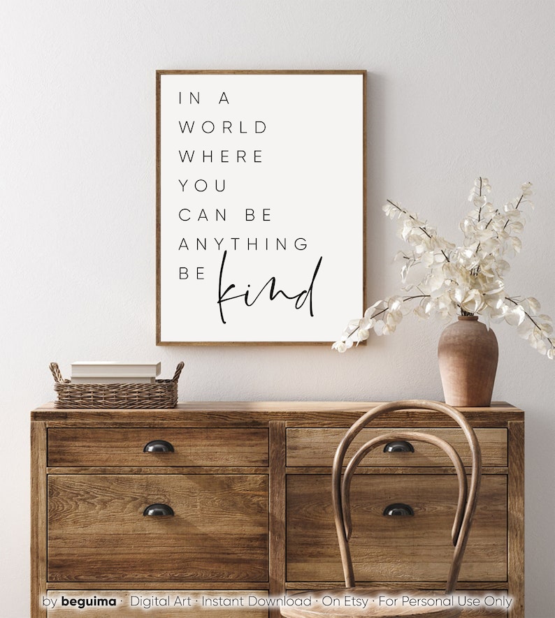 May include: A white framed print with black text that reads "In a world where you can be anything be kind". The print is hanging on a white wall above a wooden dresser with a wicker basket and a wooden chair.