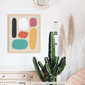 May include: A framed abstract art print featuring colorful, rounded shapes in black, white, red-orange, teal, yellow, and pink on a beige background. The artwork is displayed on a white wall, alongside a cactus and decorative elements.