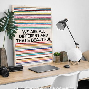 May include: A framed art print with a light wood frame. The artwork features horizontal stripes in various colours above the text "WE ARE ALL DIFFERENT AND THAT'S BEAUTIFUL" in black. The print is on a desk with a laptop, lamp, and other decor.