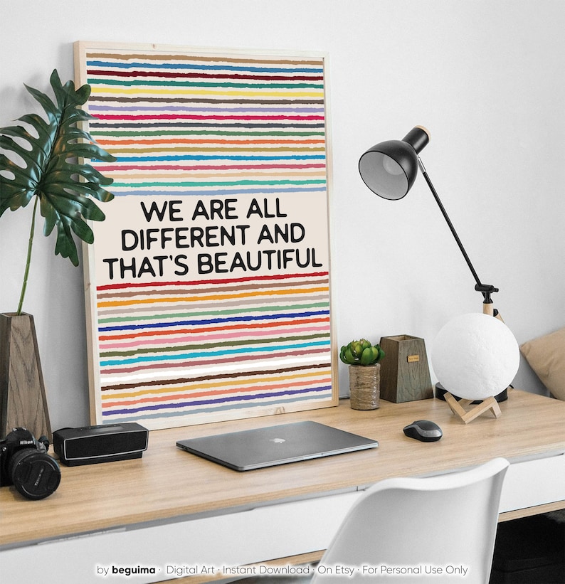 May include: A framed art print with a light wood frame. The artwork features horizontal stripes in various colours above the text "WE ARE ALL DIFFERENT AND THAT'S BEAUTIFUL" in black. The print is on a desk with a laptop, lamp, and other decor.