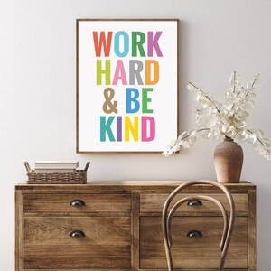 May include: A colorful printable wall art print with the text "WORK HARD & BE KIND" in a white frame. The print is on a wooden dresser with a vase of white flowers.