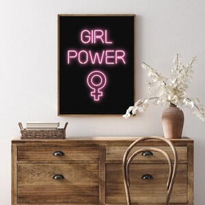 May include: A framed art print with the words "GIRL POWER" in pink neon against a black background, with the female symbol below. The print is displayed on a wooden dresser. A vase with white flowers and a wooden chair are also visible.