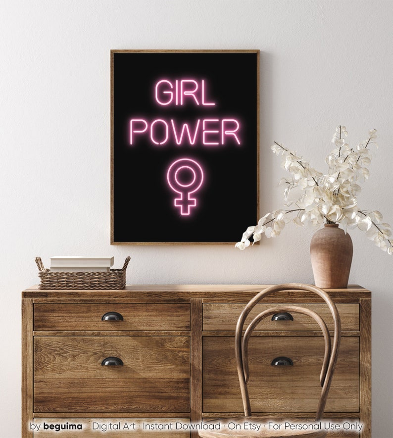 May include: A framed art print with the words "GIRL POWER" in pink neon against a black background, with the female symbol below. The print is displayed on a wooden dresser. A vase with white flowers and a wooden chair are also visible.