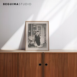 Peut inclure: Une impression encadr&eacute;e en noir et blanc d'une femme en robe longue dans une biblioth&egrave;que. L'impression est dans un cadre dor&eacute; et est expos&eacute;e sur un meuble en bois. Le texte "BEGUIMA STUDIO" est en haut.