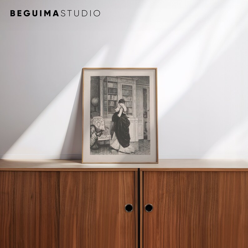 Peut inclure: Une impression encadr&eacute;e en noir et blanc d'une femme en robe longue dans une biblioth&egrave;que. L'impression est dans un cadre dor&eacute; et est expos&eacute;e sur un meuble en bois. Le texte "BEGUIMA STUDIO" est en haut.