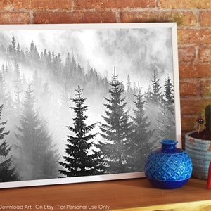 May include: Black and white photograph of a misty forest with tall pine trees. The image is framed in a white frame and is displayed on a wooden surface with a blue vase and a red wooden horse.