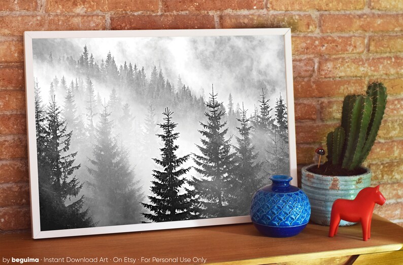 May include: Black and white photograph of a misty forest with tall pine trees. The image is framed in a white frame and is displayed on a wooden surface with a blue vase and a red wooden horse.