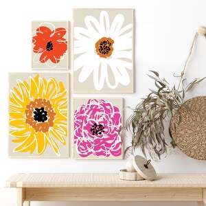 May include: Four framed floral art prints with abstract flower designs in red, white, yellow, and pink. The prints are displayed on a light beige wall. The bottom of the image shows a wooden bench with a woven top.