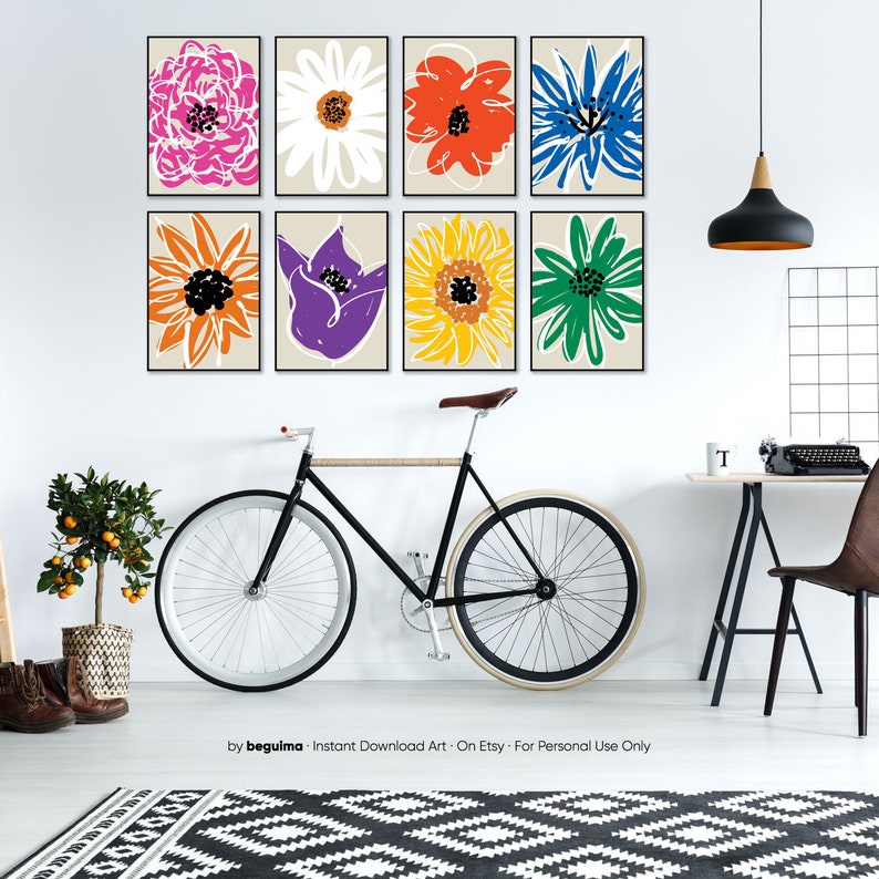 May include: Eight colorful flower illustrations in black frames on a white wall. The illustrations are of a pink flower, a white flower, a red flower, a blue flower, an orange flower, a purple flower, a yellow flower, and a green flower. The illustrations are arranged in two rows of four. The wall is in a room with a bicycle, a plant, a desk, and a chair.