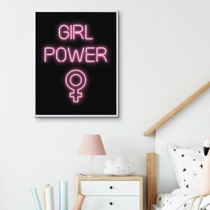 May include: A framed art print featuring the words "GIRL POWER" in pink neon-style lettering on a black background, with a pink Venus symbol below. The print is displayed on a white wall above a bedside table.