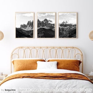 May include: Three framed black and white mountain landscape prints. The artwork features dramatic peaks, valleys, and clouds. The frames are a light wood color, and the prints are displayed above a bed with orange and white bedding.