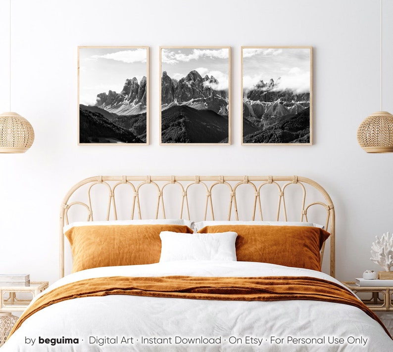 May include: Three framed black and white mountain landscape prints. The artwork features dramatic peaks, valleys, and clouds. The frames are a light wood color, and the prints are displayed above a bed with orange and white bedding.