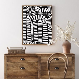 May include: Abstract black and white art print in a wooden frame. The artwork features a geometric design with vertical and curved lines. The print is displayed above a wooden dresser with a wicker basket and a vase of white flowers.