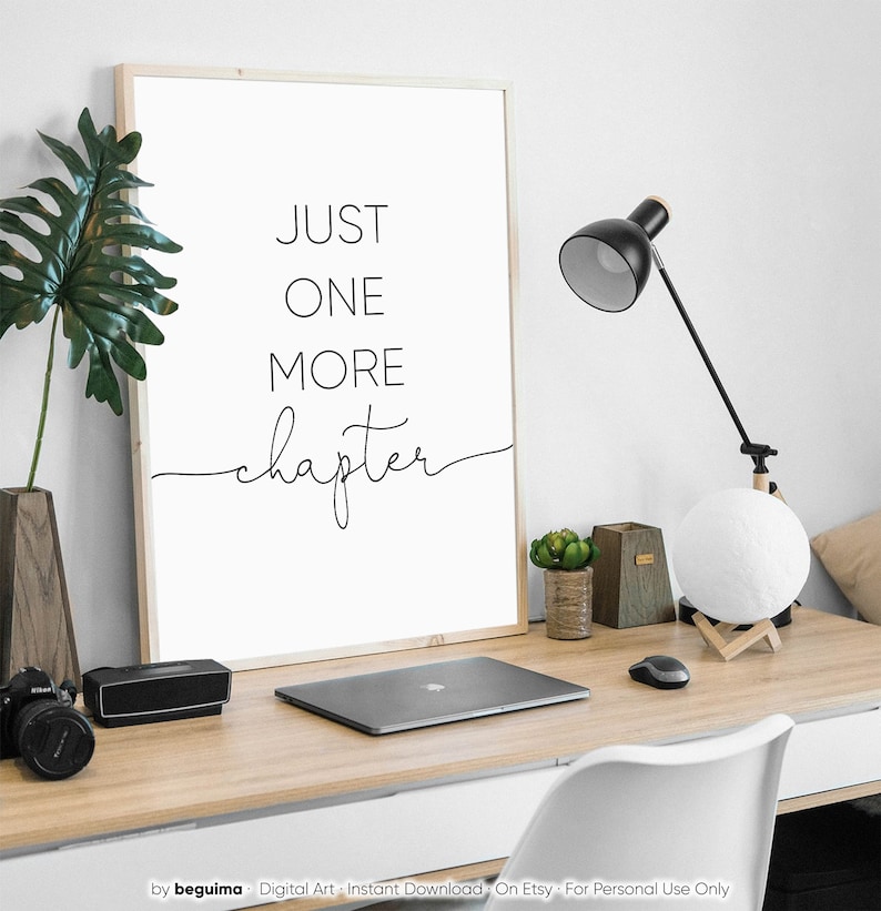 Peut inclure: Une impression murale d'art imprimable en noir et blanc avec le texte "JUST ONE MORE chapter" dans une police d'&eacute;criture de calligraphie moderne. L'impression est encadr&eacute;e et affich&eacute;e sur un bureau avec un ordinateur portable, une lampe et d'autres fournitures de bureau.