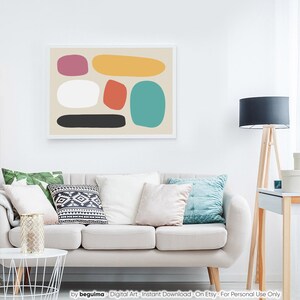 May include: Abstract art print featuring colorful, rounded shapes in pink, yellow, red, teal, white, and black against a beige background. The artwork is framed in a white border and hangs above a sofa with decorative pillows.