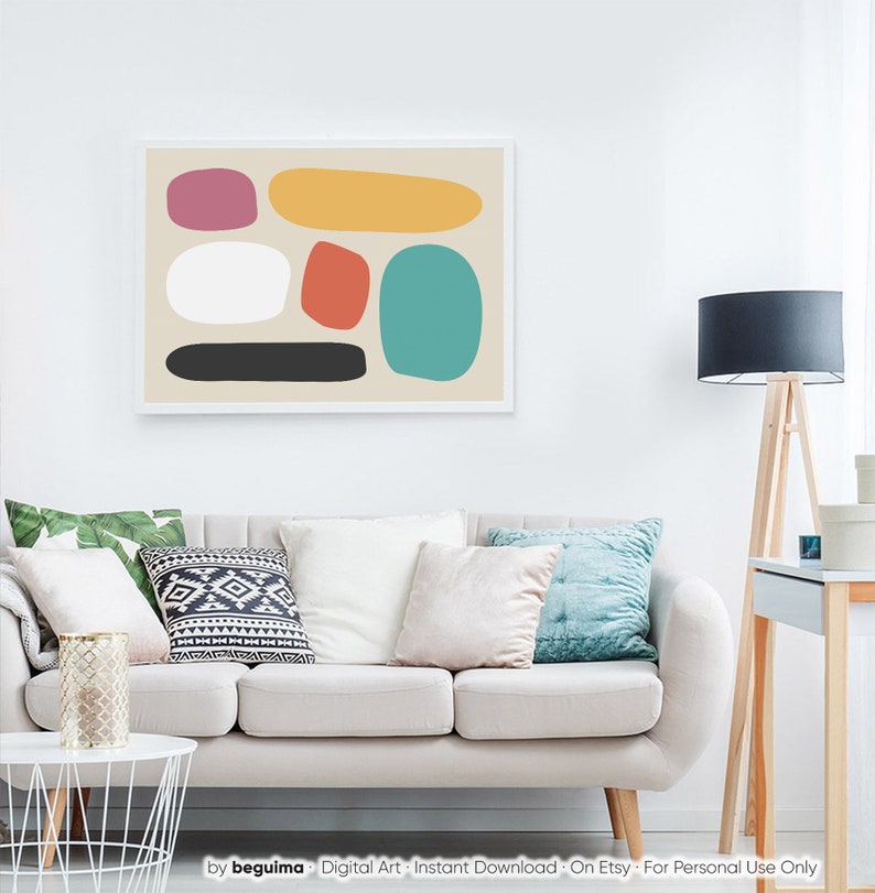 May include: Abstract art print featuring colorful, rounded shapes in pink, yellow, red, teal, white, and black against a beige background. The artwork is framed in a white border and hangs above a sofa with decorative pillows.