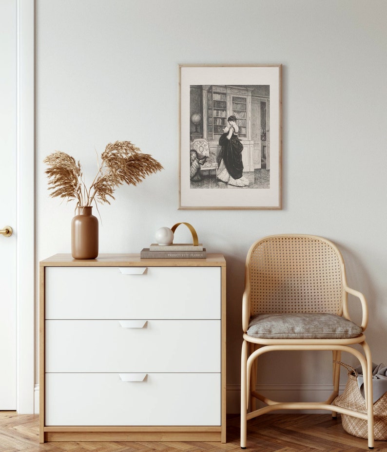 Peut inclure: Une impression encadr&eacute;e en noir et blanc d'une femme en robe longue, debout dans une biblioth&egrave;que. L'impression est suspendue au-dessus d'une commode blanche &agrave; trois tiroirs avec un dessus en bois. Un vase marron avec des plantes s&eacute;ch&eacute;es et une chaise en osier sont &eacute;galement dans l'image.