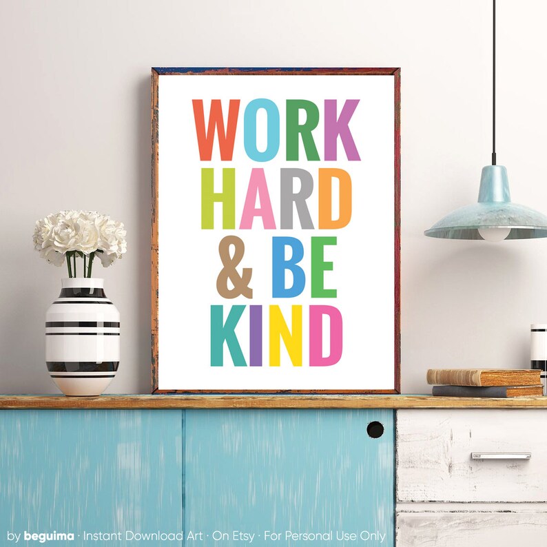 May include: A colorful printable wall art print with the text "WORK HARD & BE KIND" in a rainbow of colors. The print is framed in a brown wood frame and is hanging on a wall.