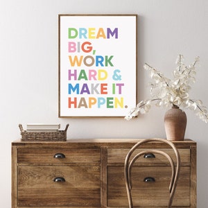 May include: A motivational quote in a colorful print reads "Dream Big, Work Hard & Make It Happen." The print is framed and hanging on a wall above a wooden dresser.