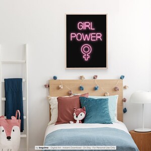 May include: A framed print with the words "GIRL POWER" in pink neon lettering, above a pink female symbol, against a black background. The print hangs above a bed with pillows and a stuffed fox. A white ladder and a fox-themed storage bag are visible.